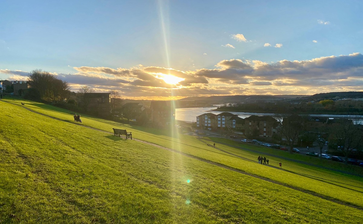A view over Churchfields in late Autumn, showing the setting sun through clouds and the steep sloping grass running down to the Esplanade and the River